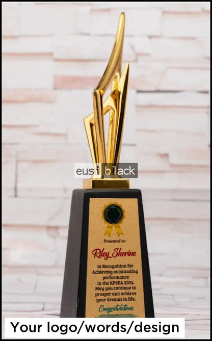 A custom-designed trophy with a polished gold abstract blade-like sculpture on a black rectangular base, featuring a gold plaque with a black seal and personalized text, set against a light brick wall background.