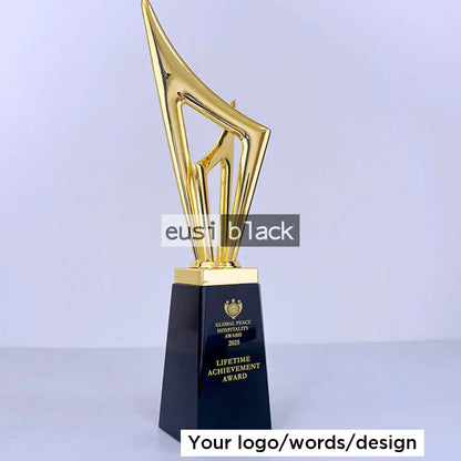 A modern gold trophy with an abstract, sweeping sculptural top mounted on a tall black pedestal base with gold lettering, photographed against a plain light gray studio background.